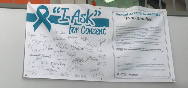 Photo and the  "I ASK" for consent banner containing signatures of Virginia Tech students displayed in Squires Student Center. 